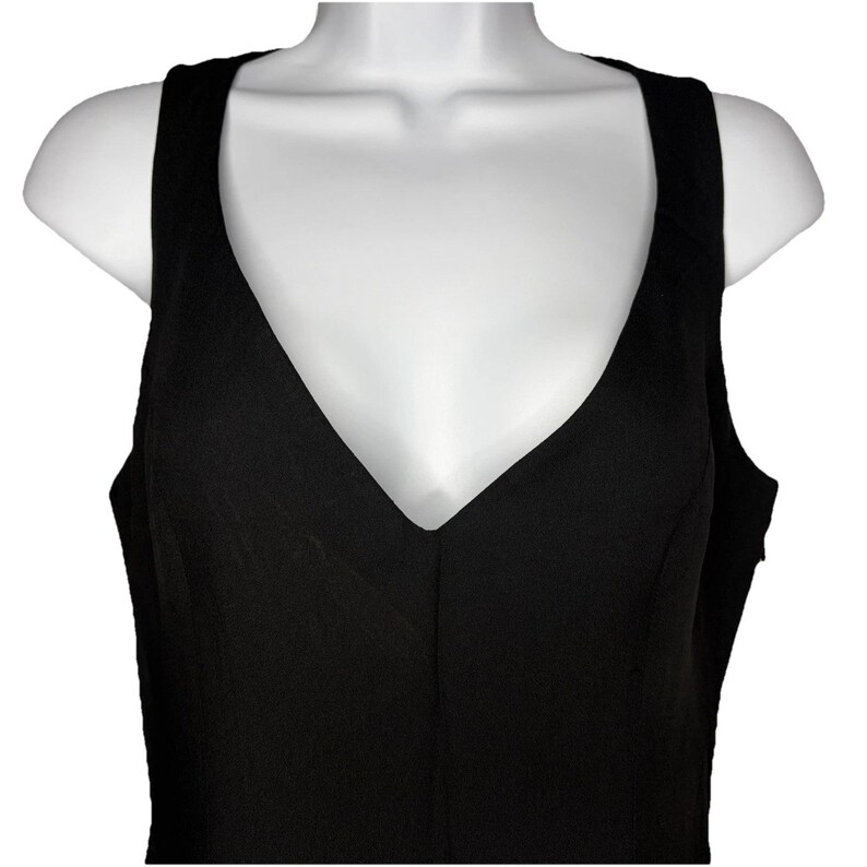 Op de afbeelding: Een zwarte mouwloze jurk met een diepe V-hals. De jurk wordt getoond op een witte mannequin torso, waarbij de snit en stijl worden benadrukt. De stof heeft een effen, matte uitstraling.