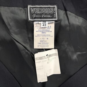 Op de afbeelding: Close-up van de binnenkant van een zwart kledingstuk, met labels van het merk Versus Gianni Versace. De labels tonen tekst in het Italiaans en Japans, met details over de stofsamenstelling en de kledingstijl.