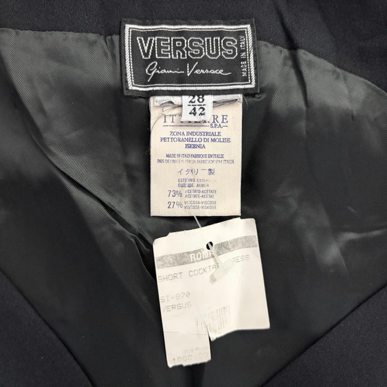 Op de afbeelding: Close-up van de binnenkant van een zwart kledingstuk, met labels van het merk Versus Gianni Versace. De labels tonen tekst in het Italiaans en Japans, met details over de stofsamenstelling en de kledingstijl.