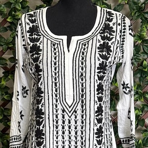 May include: White long dress with black floral embroidery. The dress features a V-neckline and long sleeves. The embroidery is detailed and covers the front, sleeves, and hem of the dress. The dress is displayed on a black mannequin.
