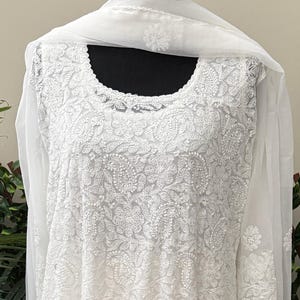 Könnte beinhalten: Ein weißes, besticktes Tunika- und Schal-Set. Die Tunika hat detaillierte Blumen- und Paisley-Muster, lange Ärmel und einen runden Ausschnitt. Der transparente Schal ist über die Schultern drapiert. Das Outfit wird auf einer Schaufensterpuppe präsentiert.