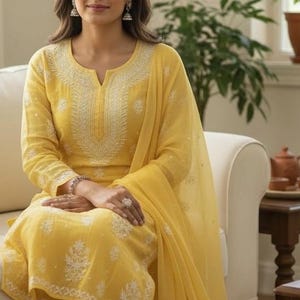 May include: A woman wearing a yellow embroidered salwar kameez, a traditional South Asian outfit, sits on a white sofa. The outfit includes a tunic, trousers, and a scarf, all in a bright yellow hue with white floral embroidery. The background includes a window and plants.