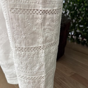 May include: Close-up of white pants with intricate floral embroidery and cutwork details. The fabric appears to be cotton or a similar lightweight material, ideal for warm weather. The design features detailed stitching and openwork patterns.