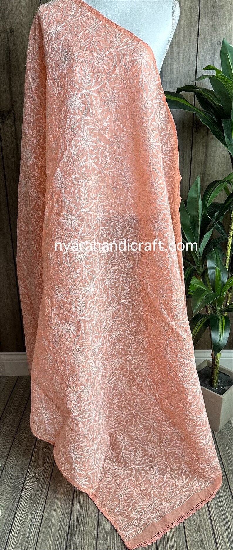 May include: A peach-colored sheer fabric with white floral embroidery. The fabric has a crocheted edge and the text "nyarahandicraft.com" is visible in the lower center of the image.
