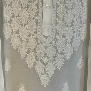 May include: White sheer tunic with white embroidered floral design. The tunic has a button-up collar and a long sleeve.