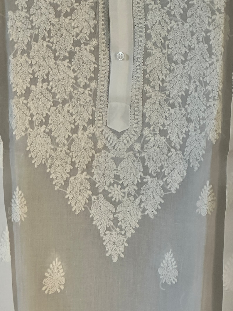 May include: White sheer tunic with white embroidered floral design. The tunic has a button-up collar and a long sleeve.