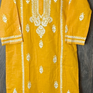 Könnte beinhalten: Eine gelbe Baumwolltunika mit weißen Stickereien. Die Stickerei zeigt Blumenmuster und ein Paisley-Muster. Die Tunika hat einen V-Ausschnitt und 3/4 Ärmel.