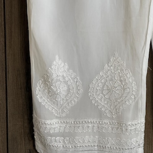 May include: White pants with intricate, embroidered floral patterns near the hem. The embroidery is detailed and adds a decorative element to the garment. The pants are made of a lightweight fabric.