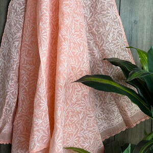 May include: A peach-colored sheer fabric with a white floral embroidery pattern. The fabric is draped over a wooden surface and has a delicate lace trim along the edge.