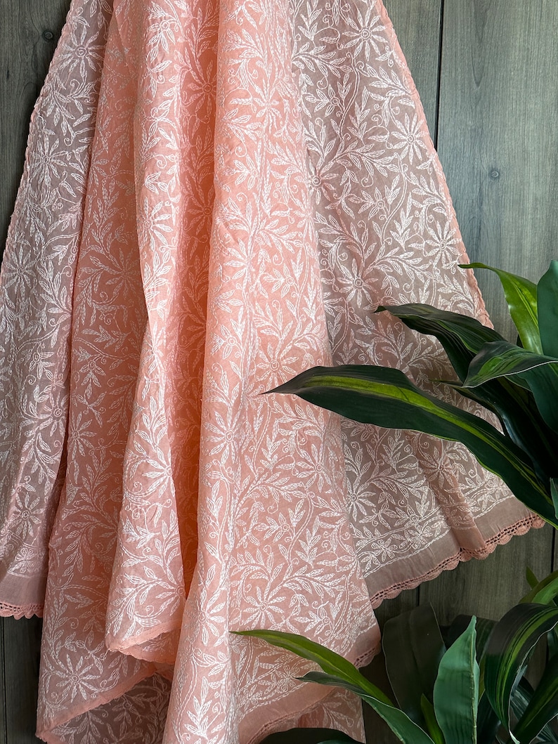 May include: A peach-colored sheer fabric with a white floral embroidery pattern. The fabric is draped over a wooden surface and has a delicate lace trim along the edge.
