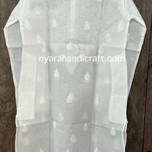 May include: White cotton tunic with a delicate embroidered pattern of white leaves. The tunic has a long, flowing silhouette and a stand-up collar. The text "nyarahandicraft.com" is printed on the front of the tunic.