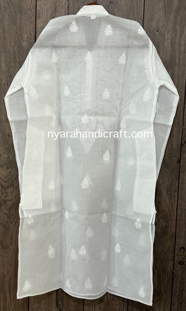 May include: White cotton tunic with a delicate embroidered pattern of white leaves. The tunic has a long, flowing silhouette and a stand-up collar. The text "nyarahandicraft.com" is printed on the front of the tunic.