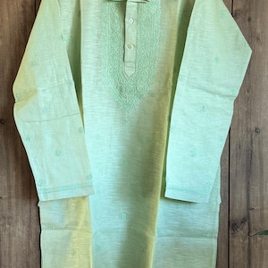 May include: A light green tunic with long sleeves and a mandarin collar. The tunic features intricate embroidered detailing around the collar and down the front, with small floral embroidery scattered across the fabric. The garment is made of a lightweight material.