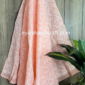 May include: A peach-colored sheer fabric shawl with white floral embroidery. The shawl has a delicate lace trim and is draped over a wooden surface.