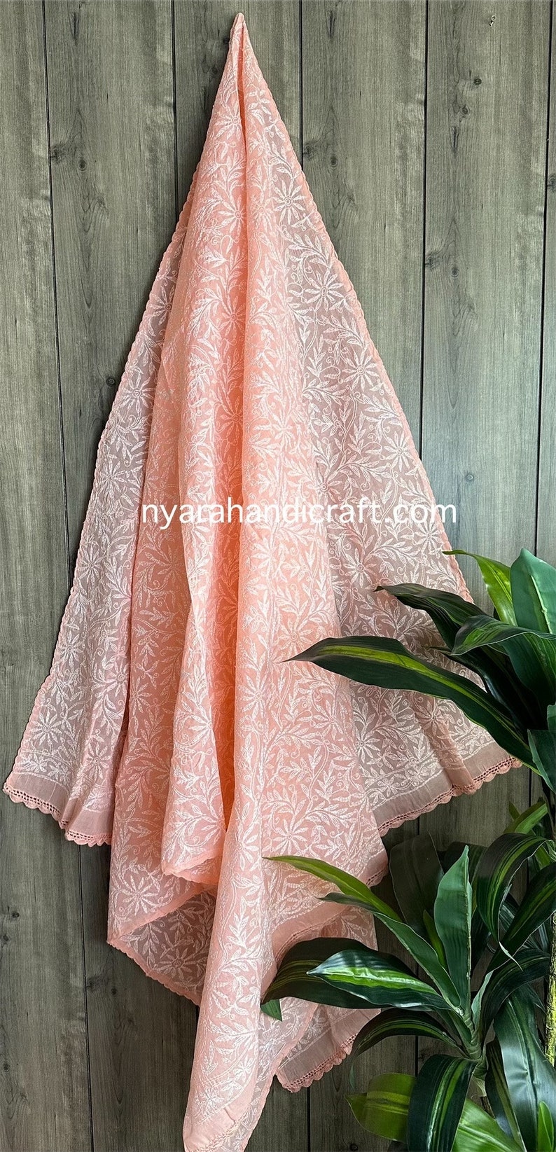 May include: A peach-colored sheer fabric shawl with white floral embroidery. The shawl has a delicate lace trim and is draped over a wooden surface.