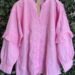 May include: A light pink, long-sleeved blouse with intricate embroidered floral patterns. The blouse features a mandarin collar, a button-down front, and ruffled detailing on the sleeves. The fabric appears lightweight and airy, suitable for warmer weather.