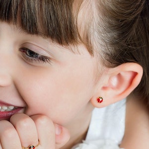 May include: A young girl with brown hair wearing a red and gold ladybug stud earring.