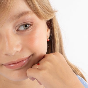 May include: A gold-colored ring and earrings set featuring a red ladybug design. The ring is on a finger, and the earrings are dangling from the ear. The ladybug design has black spots and a red body.