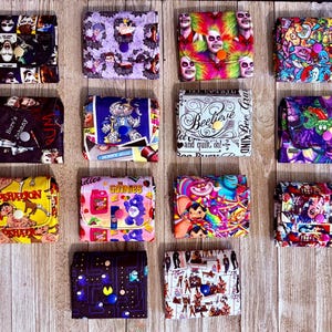 May include: Fourteen square fabric wallets with snap closures, each featuring a unique colorful pattern including movie characters, cartoon themes, and graphic prints. The wallets are arranged in a grid, showcasing various designs and vibrant colors.