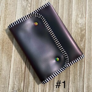 May include: A dark gray clutch with a black and white striped trim and iridescent snap closures. The clutch is rectangular and has a folded flap. The number "#1" is printed in black on the bottom right corner.