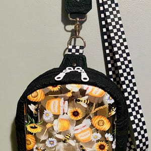 May include: A black crossbody bag with a clear front panel featuring a repeating pattern of honey jars, bees, sunflowers, and daisies. The bag has a black and white checkered strap.