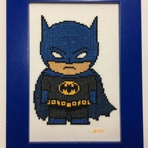 May include: Cross-stitch artwork of Batman in a blue frame. The character is depicted in a pixelated style, wearing a blue cowl and cape, a black suit with a yellow bat symbol, and the text "JOKO ODOL" on the belt. The artwork is signed "AN17".