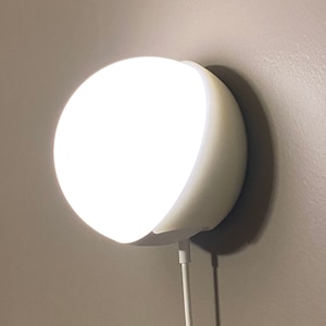 Puede incluir: Una luz nocturna redonda blanca enchufada a una toma de corriente en una pared. La luz está encendida e ilumina la pared.