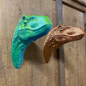 May include: Two dinosaur head sculptures mounted on a wooden wall. One is a bright green color and the other is a copper color.
