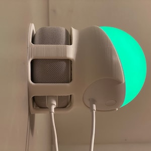 May include: White 3D printed wall mount for a smart speaker and a smart light bulb. The speaker is in the mount and the light bulb is attached to the side of the mount. Both devices are plugged in and have white cords.