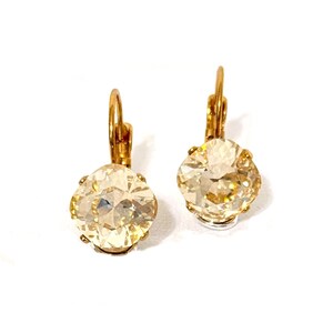 May include: A pair of gold-colored leverback earrings. Each earring features a large, round, light-yellow crystal. The crystals are faceted, catching the light. The earrings are set against a white background.