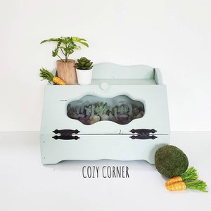 May include: A light blue wooden bread box with a glass window and black hinges. The box has a distressed finish and the words "COZY CORNER" are printed on the front. The box is decorated with small green plants and carrots.
