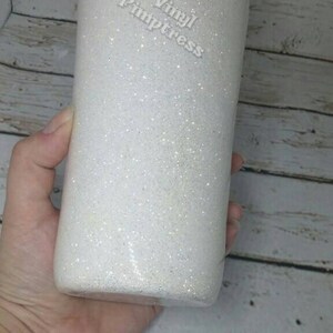 May include: A white glitter tumbler with the text "Vinyl Pimpstress" in a cursive font. The tumbler is cylindrical and appears to be held by a person. The background is a light-colored wooden surface.