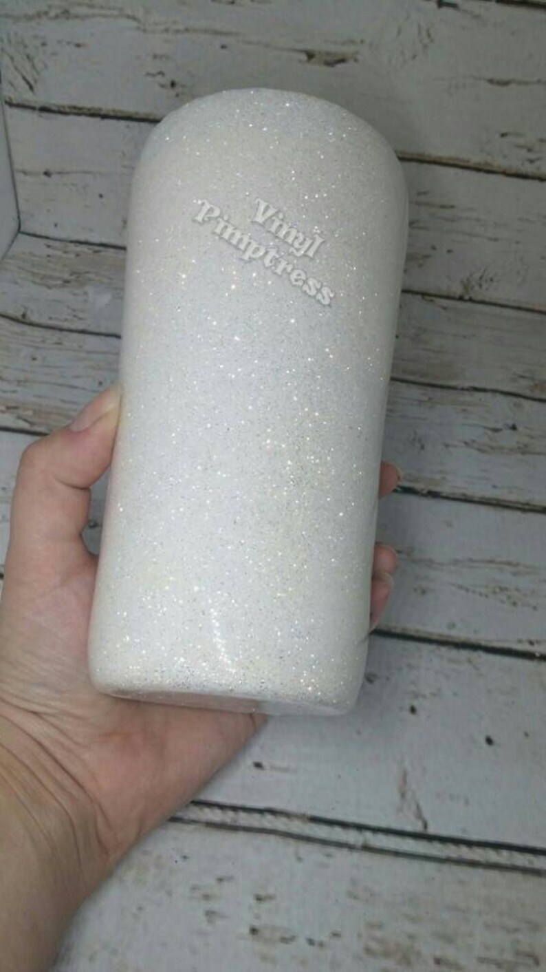 May include: A white glitter tumbler with the text "Vinyl Pimpstress" in a cursive font. The tumbler is cylindrical and appears to be held by a person. The background is a light-colored wooden surface.