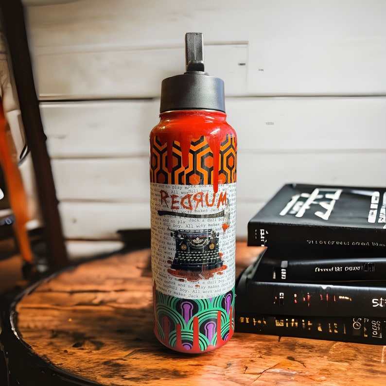Puede incluir: Una botella de agua roja con tapa negra y boquilla plateada. La botella presenta un dise&ntilde;o inspirado en una pel&iacute;cula de terror, con la palabra "REDRUM" y un gr&aacute;fico de m&aacute;quina de escribir vintage. El dise&ntilde;o incluye patrones geom&eacute;tricos y texto.