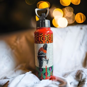 Puede incluir: Una botella de agua roja con asa y tapa negras. La botella presenta un patr&oacute;n geom&eacute;trico repetitivo en naranja y negro, un collage de texto y un gr&aacute;fico de una persona. La parte inferior tiene un dise&ntilde;o verde y morado.