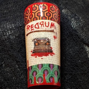 Puede incluir: Un vaso cil&iacute;ndrico con tem&aacute;tica de pel&iacute;cula de terror. El dise&ntilde;o incluye la palabra "REDRUM" en sangre goteando, una m&aacute;quina de escribir y un hacha. El vaso tiene una parte superior e inferior rojas con un patr&oacute;n geom&eacute;trico.