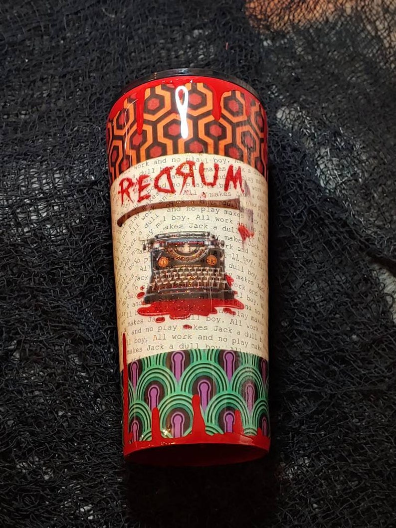 Puede incluir: Un vaso cil&iacute;ndrico con tem&aacute;tica de pel&iacute;cula de terror. El dise&ntilde;o incluye la palabra "REDRUM" en sangre goteando, una m&aacute;quina de escribir y un hacha. El vaso tiene una parte superior e inferior rojas con un patr&oacute;n geom&eacute;trico.