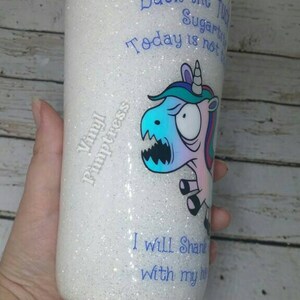 May include: A white glitter tumbler with blue text and a cartoon unicorn graphic. The text includes phrases like "Back the f..." and "I will Shank..." The tumbler is cylindrical and held by a hand.