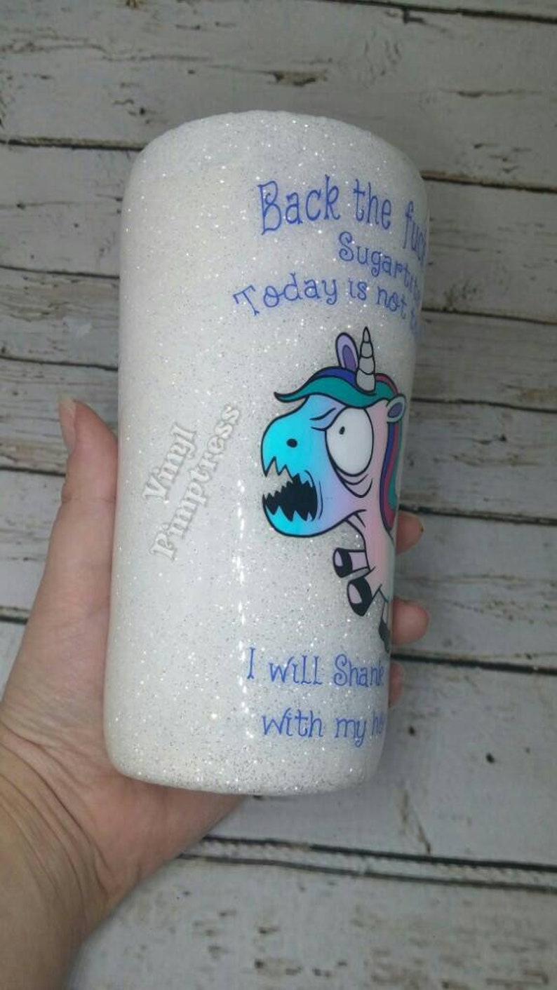May include: A white glitter tumbler with blue text and a cartoon unicorn graphic. The text includes phrases like "Back the f..." and "I will Shank..." The tumbler is cylindrical and held by a hand.