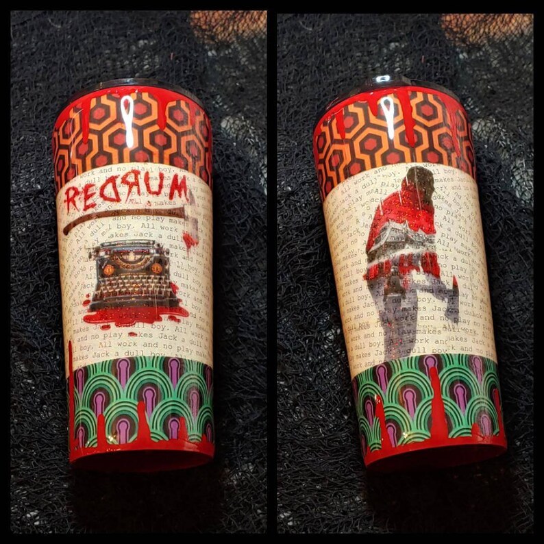 Puede incluir: Un vaso cil&iacute;ndrico con im&aacute;genes de la pel&iacute;cula "El Resplandor". El dise&ntilde;o incluye la palabra "REDRUM", una m&aacute;quina de escribir, un hacha y una escena de hotel. El vaso tiene un patr&oacute;n geom&eacute;trico rojo y naranja en la parte superior e inferior.