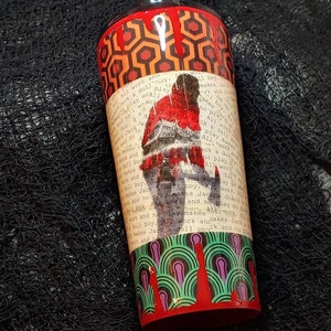 Puede incluir: Un vaso cil&iacute;ndrico con un patr&oacute;n geom&eacute;trico rojo y naranja en la parte superior e inferior. El centro presenta una imagen desgastada de una persona con el texto "All work and no play makes Jack a dull boy."