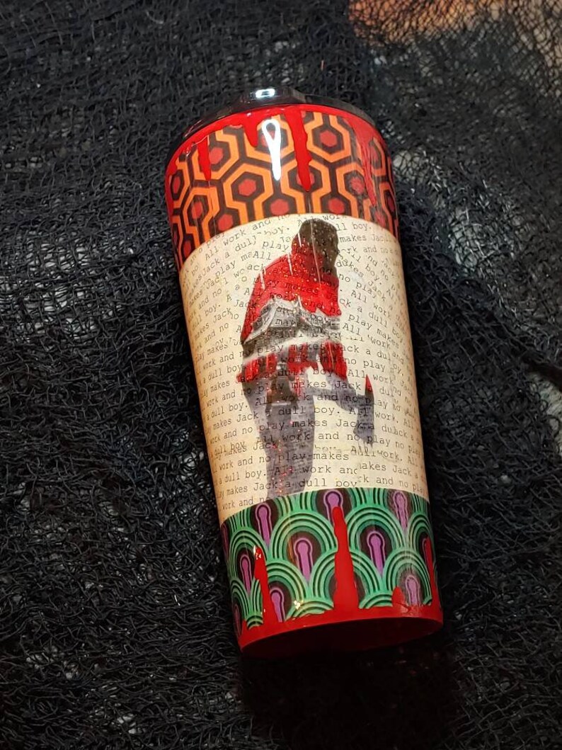 Puede incluir: Un vaso cil&iacute;ndrico con un patr&oacute;n geom&eacute;trico rojo y naranja en la parte superior e inferior. El centro presenta una imagen desgastada de una persona con el texto "All work and no play makes Jack a dull boy."