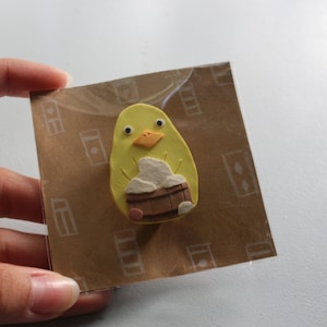 May include: A yellow chick figurine holding a small brown bucket with white cotton balls inside. The figurine is on a brown background with a white pattern.