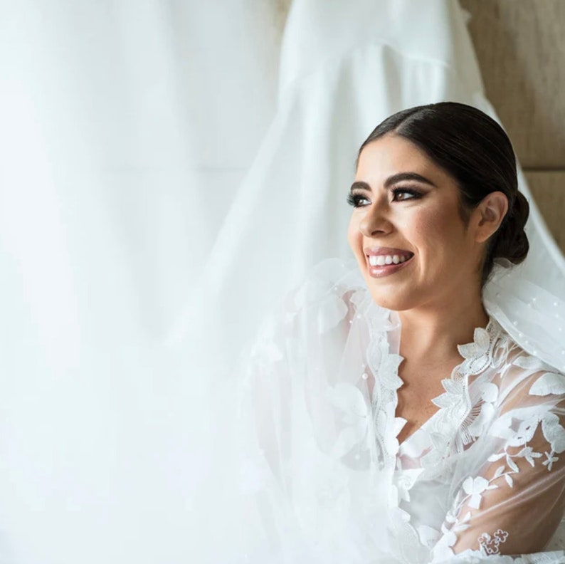 May include: A bride smiles while wearing a white lace robe and veil. She is standing in front of a white curtain.