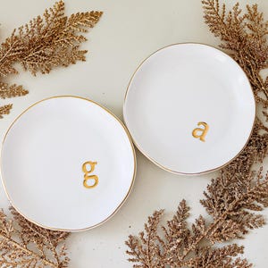 May include: Two white ceramic ring dishes with gold trim and gold lettering. The dish on the left says "g" and the dish on the right says "a".