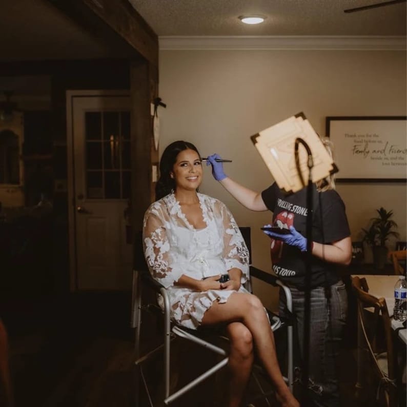 May include: A woman in a white lace robe sits in a chair while another woman applies makeup to her face. The woman applying makeup is wearing a black t-shirt with the words "Rolling Stone" on it.
