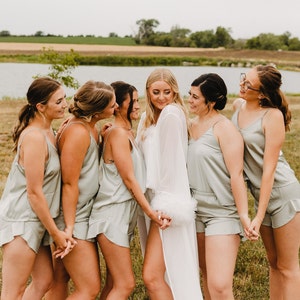 May include: A group of women in matching sage green silk pajama sets and a bride in a white robe with feather trim. They are standing outside on a grassy area near a body of water, holding hands and smiling.
