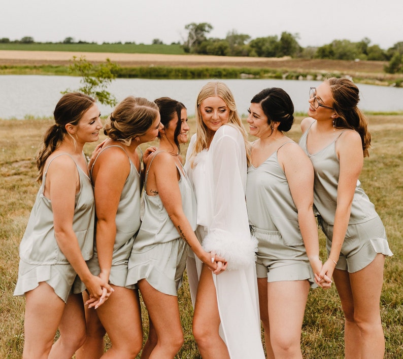 May include: A group of women in matching sage green silk pajama sets and a bride in a white robe with feather trim. They are standing outside on a grassy area near a body of water, holding hands and smiling.