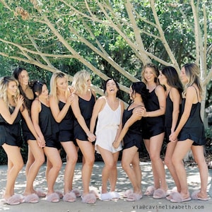 May include: A group of women wearing black satin pajama sets and pink fluffy slippers. One woman wears a white pajama set. They are standing outside under a tree, smiling and laughing.