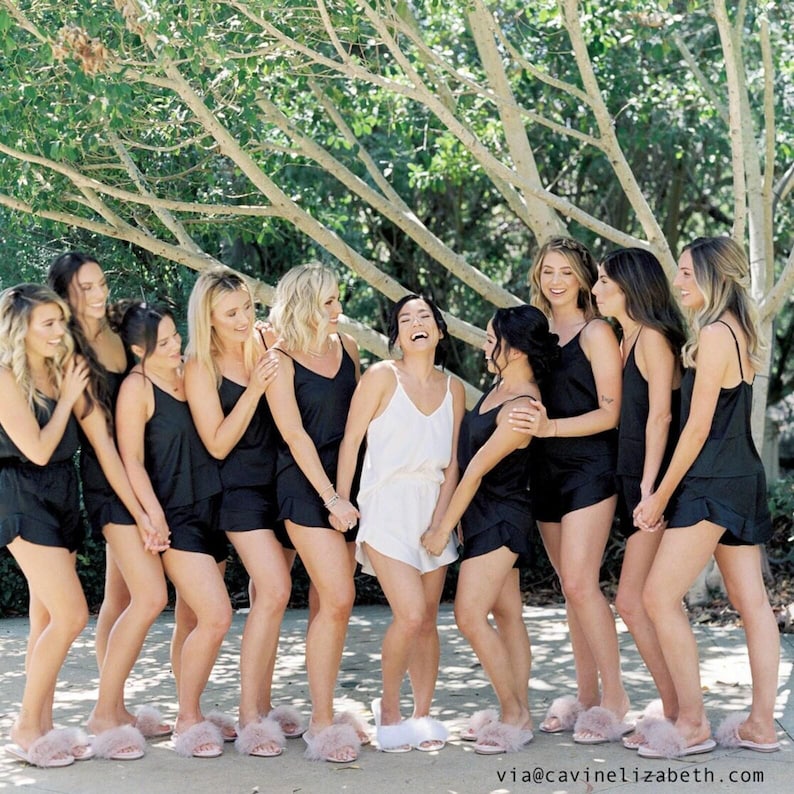 May include: A group of women wearing black satin pajama sets and pink fluffy slippers. One woman wears a white pajama set. They are standing outside under a tree, smiling and laughing.